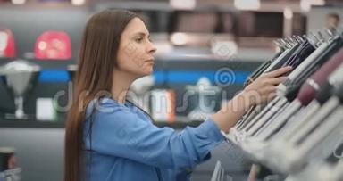 在电器商店里，一个穿着衬衫的黑发女人通过观看和拿着设备来选择搅拌机购物